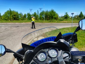 Umundurowany policjant mierzy prędkość pojazdom ręcznym miernikiem prędkości. Widoczny motocykl policyjny.