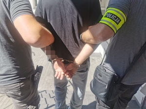 Dwóch policjantów w ubraniach cywilnych z opaską na ręku z napisem policja prowadzą zatrzymanego mężczyznę, który ma założone kajdanki na ręce trzymane z tyłu.