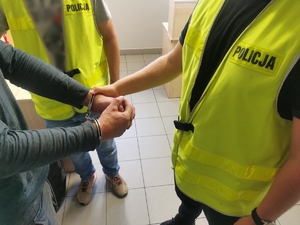 Dwóch policjantów w ubraniach cywilnych w kamizelkach z napisem policja prowadzą zatrzymanego mężczyznę, który ma założone kajdanki na ręce trzymane z przodu.