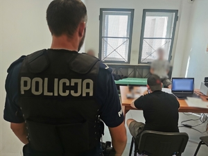 Pomieszczenie dla osób zatrzymanych, przy kracie siedzi na ławce dwóch zatrzymanych. Jeden z policjantów w ubraniu cywilnym siedzi przy biurko, a drugi umundurowany stoi.