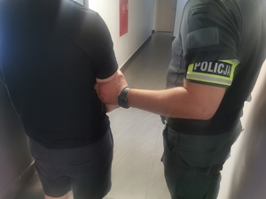 Policjant w ubraniu cywilnym z opaską na ręku z napisem policja trzyma zatrzymanego mężczyznę, który ma założone kajdanki na ręce trzymane z przodu.
