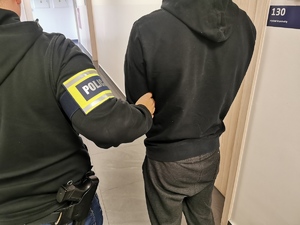 Policjant w ubraniu cywilnym z opaską na ręku z napisem policja trzyma zatrzymanego mężczyznę na korytarzu komendy