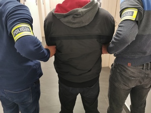Dwóch policjantów w ubraniu cywilnym z opaską na ręku z napisem policja trzyma zatrzymanego mężczyznę