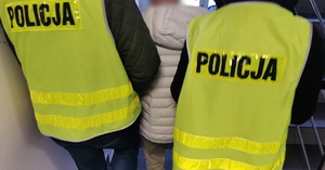 Dwóch policjantów w ubraniach cywilnych, w kamizelkach odblaskowych z napisem policja prowadzi po schodach zatrzymanego mężczyznę.