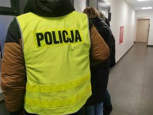 Policjant w ubraniu cywilnym, w kamizelce z napisem policja trzyma zatrzymaną kobietę na korytarzu komendy.
