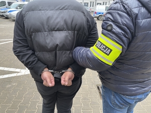 Policjant w ubraniu cywilnym z opaską na ręku z napisem policja prowadzi zatrzymanego mężczyznę